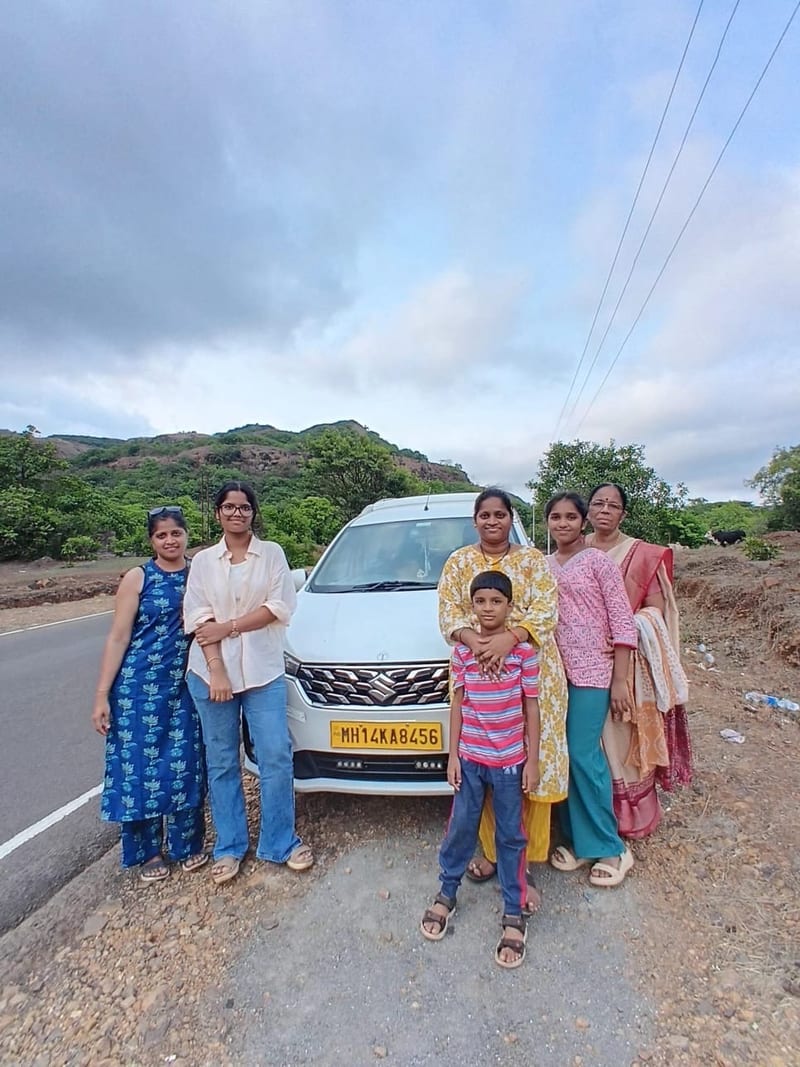 Group of six travellers posing beside a white car on a rural road with hills behind