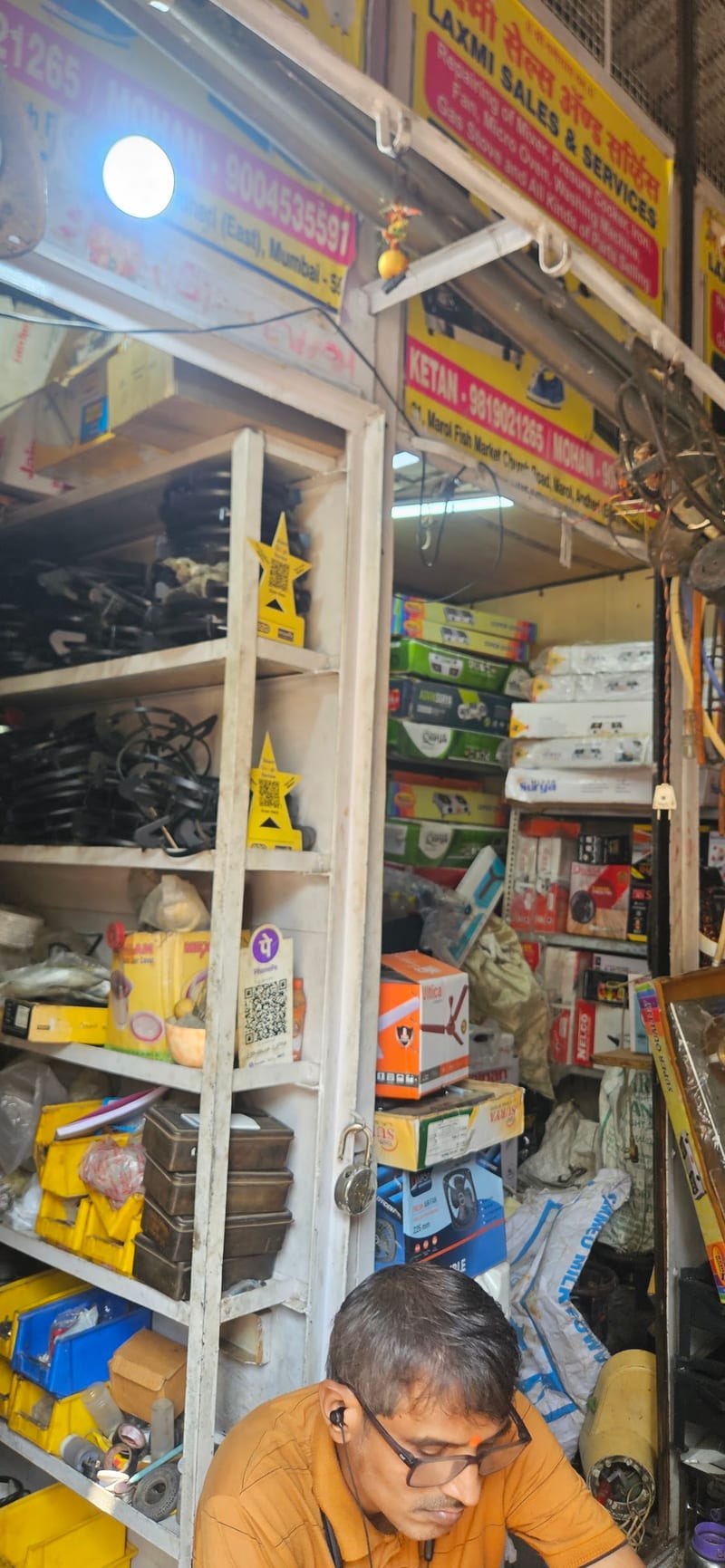 Narrow shop interior with shelves of boxed goods and a seated man near the entrance