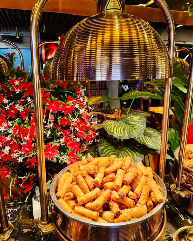 Fried spring rolls on a buffet tray beneath a heat lamp with floral decor