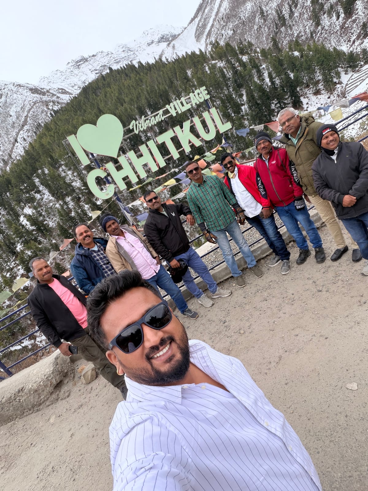Travellers posing at a Chitkul sign with snow-covered mountains behind