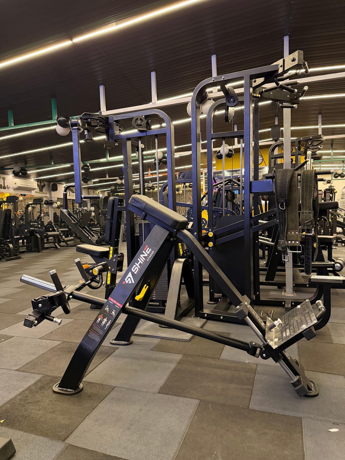 Strength-training and exercise machines lined up under linear lighting in a gym