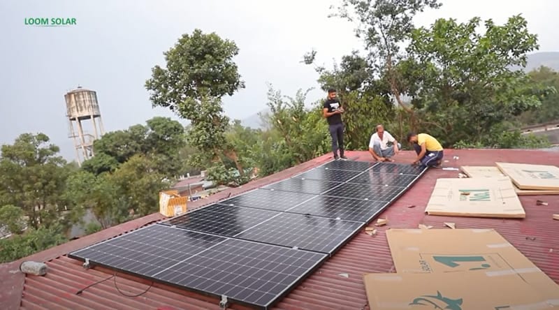 Workers installing solar panels on a red rooftop surrounded by trees