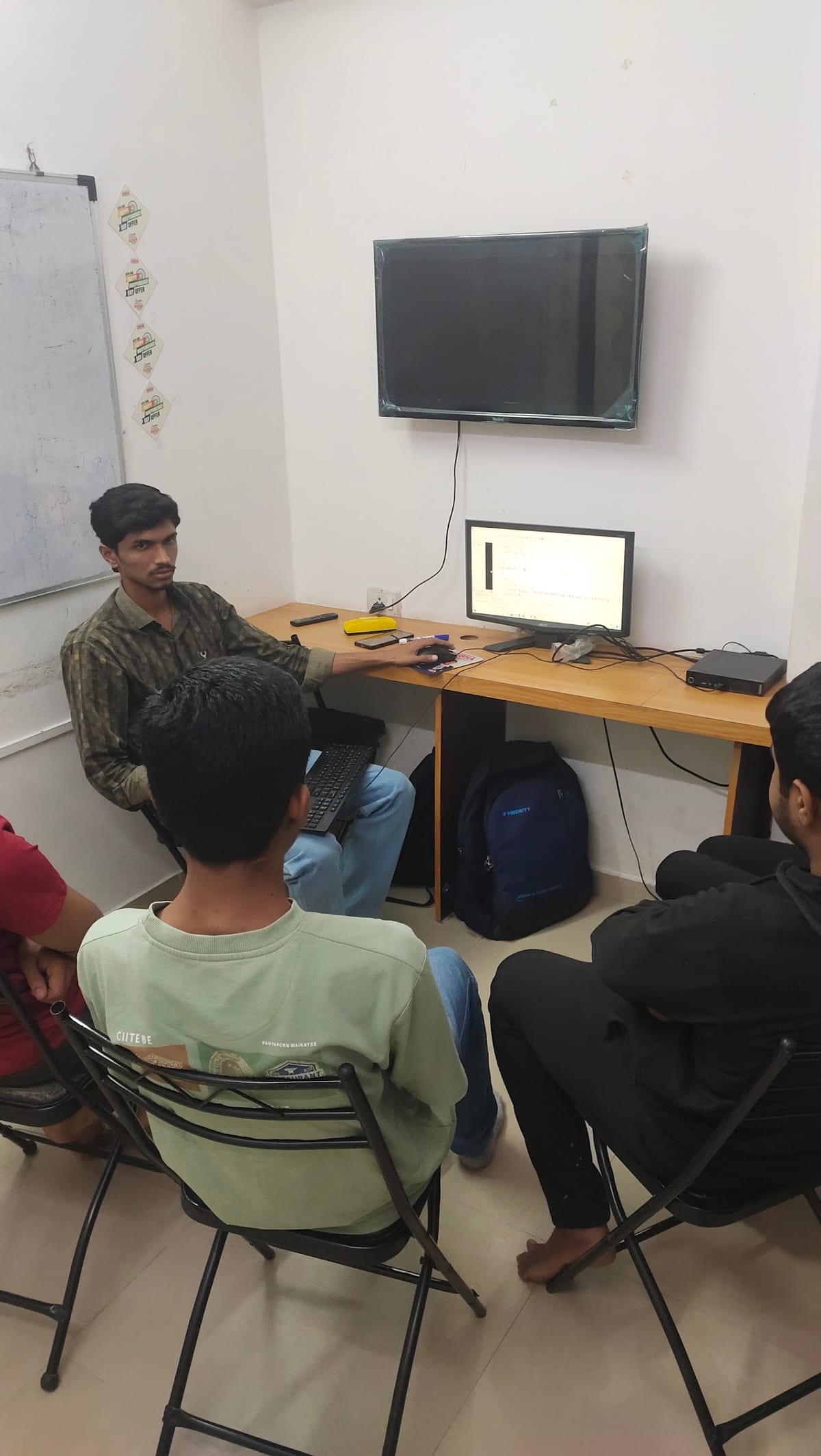 Classroom with seated learners, a wall-mounted television, computer monitor, desk and whiteboard