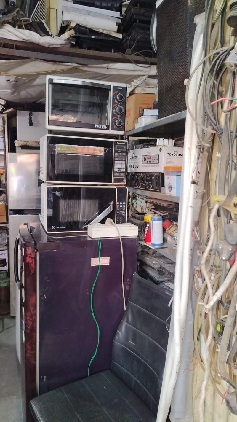 Crowded repair shop interior with stacked ovens, appliances, tools and bundled cables
