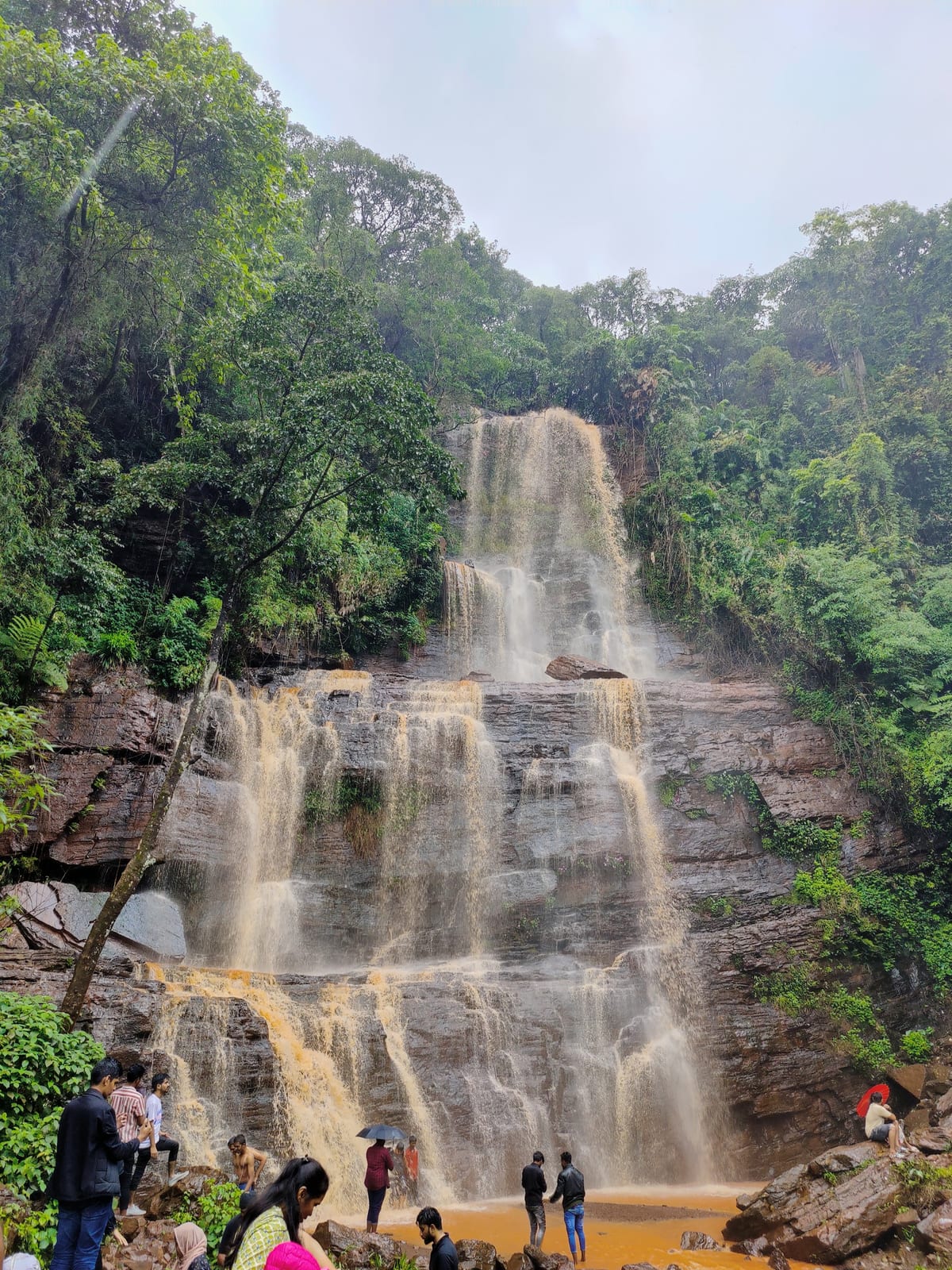 Tall waterfall over layered rock in a forest with people at its base