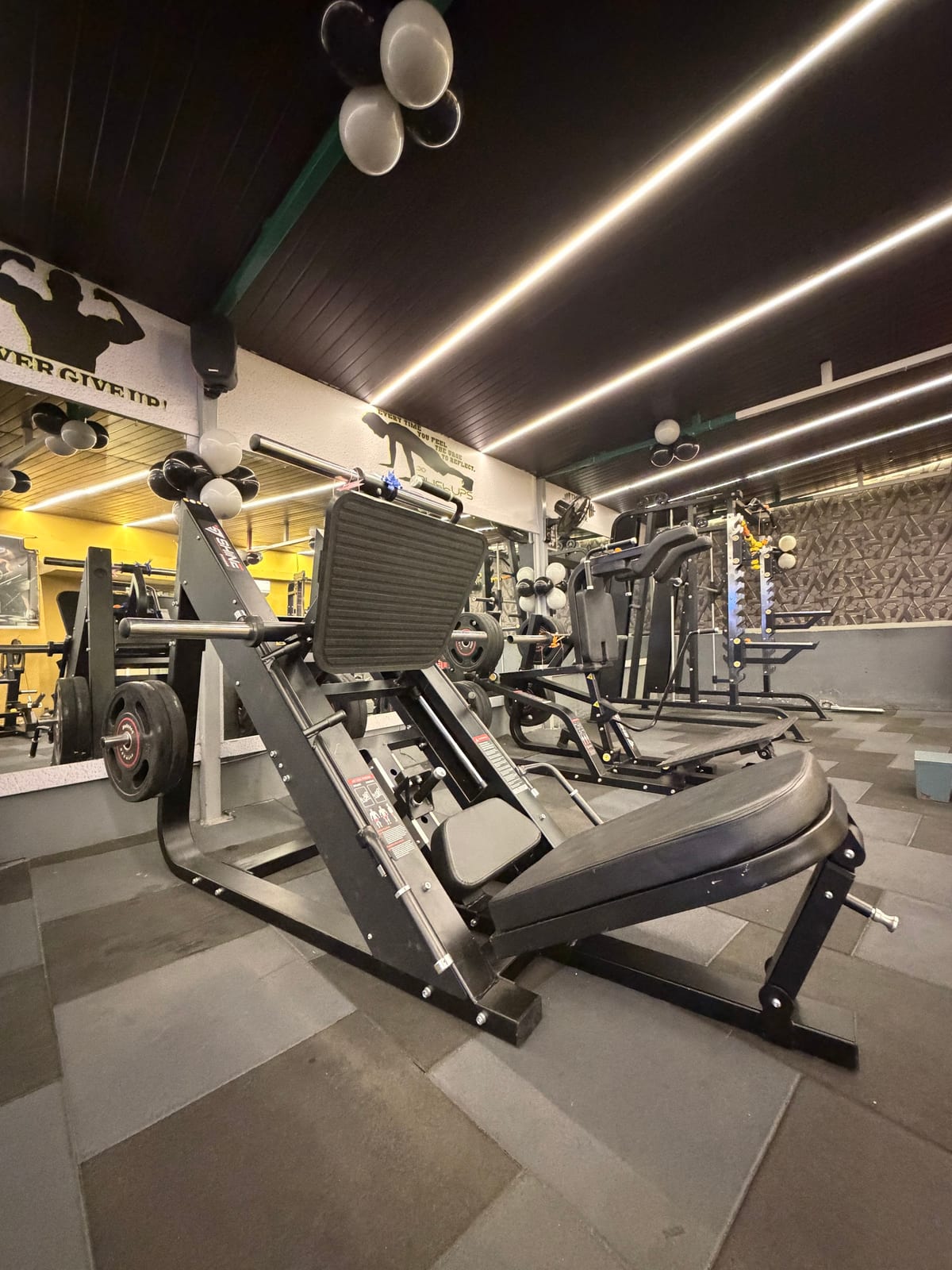 Multiple strength-training machines and weights on the gym floor beneath bright ceiling lighting