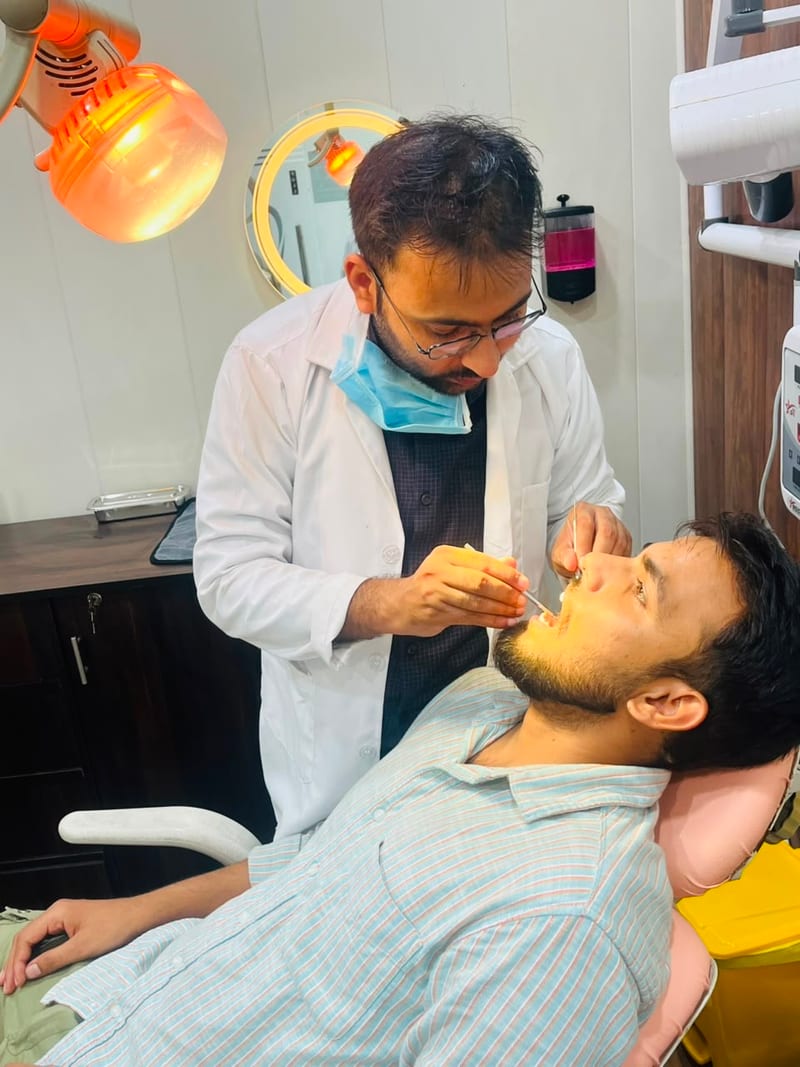Practitioner in a white coat examining a reclining man's mouth under a dental lamp