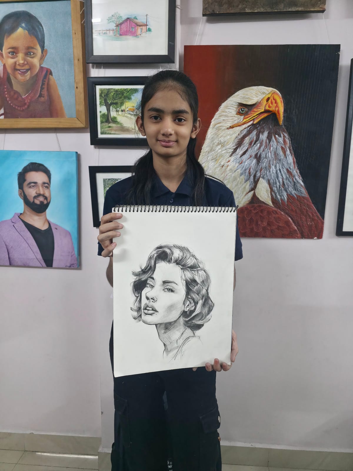 Young person holding a pencil portrait drawing in front of framed artwork including an eagle painting
