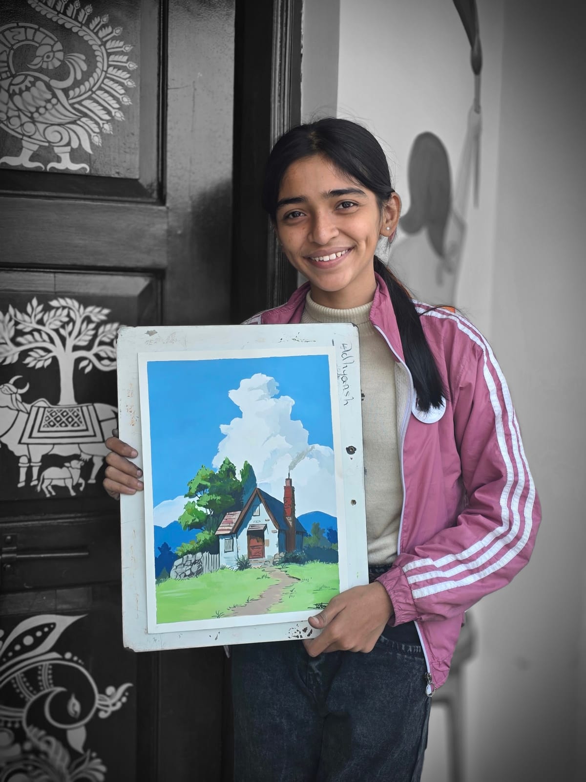 Smiling girl in a pink jacket holding a framed landscape painting by a carved wooden door