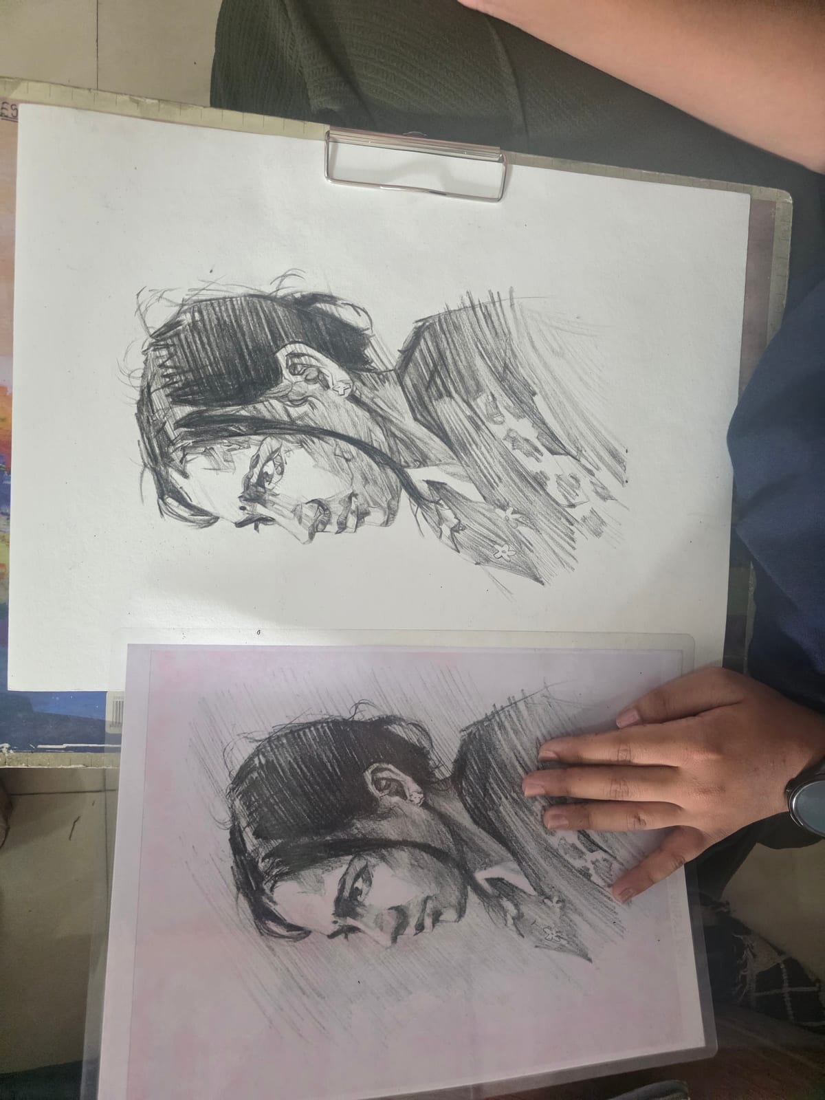 Two similar portrait sketches on paper with a hand resting on the lower drawing